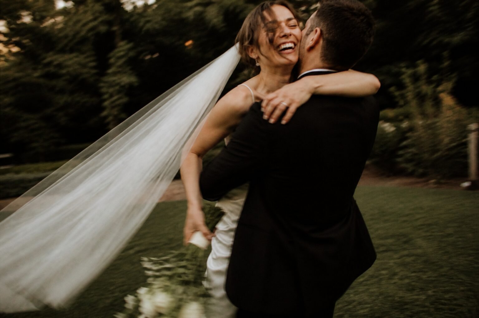 Bride laughing as she is lifted beneath dark woodland trees