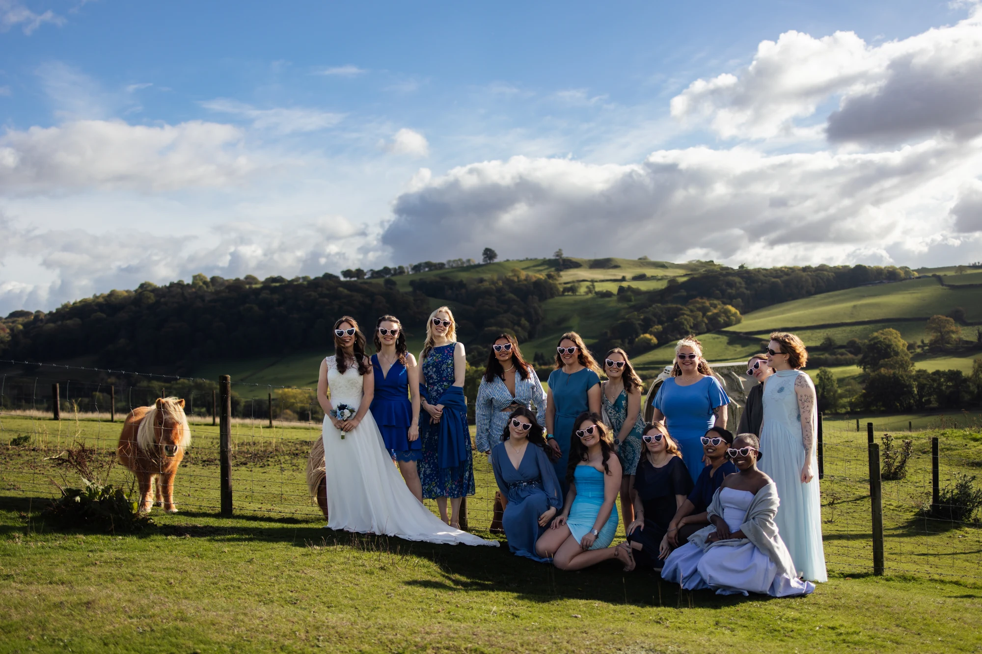 The bride and friends wearing heart-shaped sunglasses, with a pony and green hills behind them