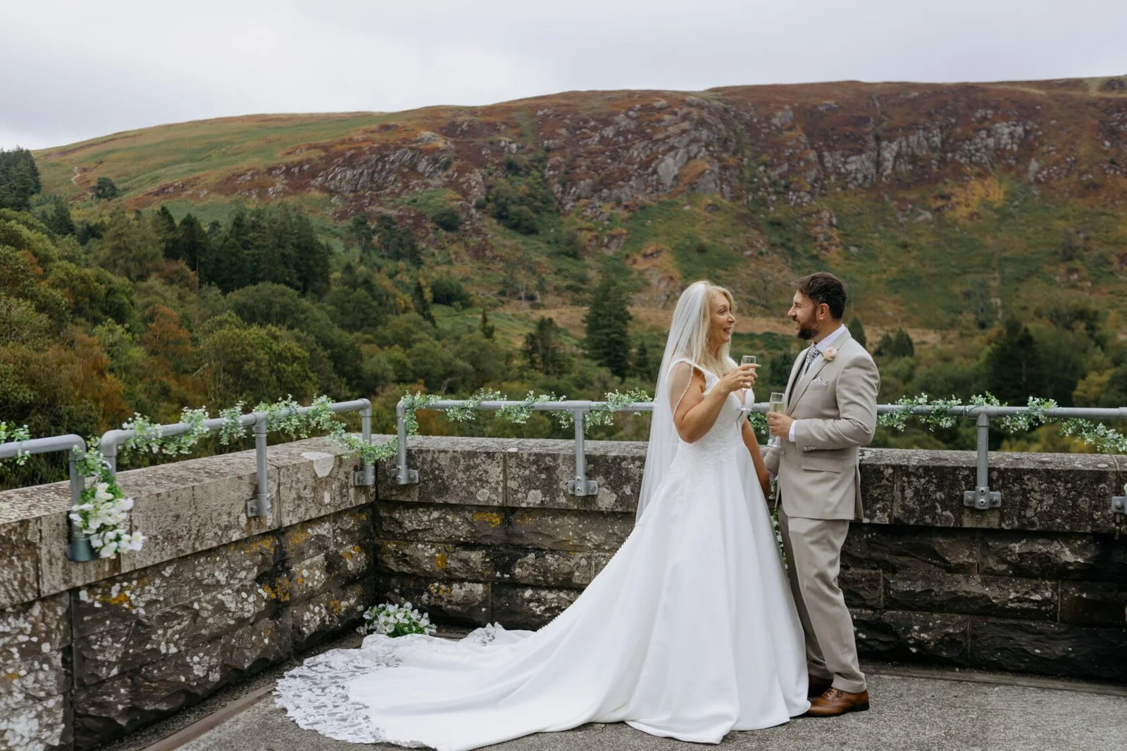 Newlyweds holding champagne on a stone terrace overlooking the Elan Valley