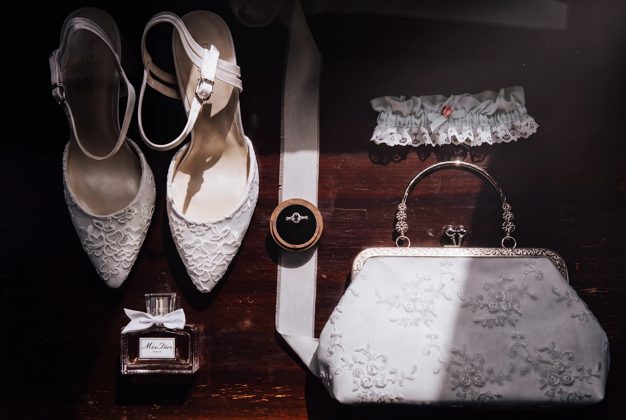Lace bridal shoes, a ring, perfume, garter and embroidered handbag arranged on dark wood